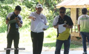 Three men standing outdoors, reading papers; the man in white points toward something.